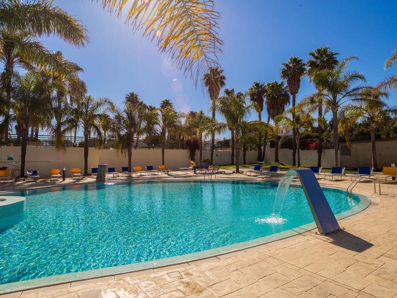 Großer, blauer Pool mit Palmen rundherum und Sonnenliegen auf sandfarbenem Boden.