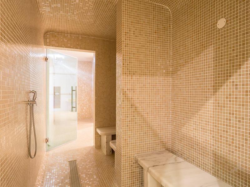 Moderner Dampfraum mit kleinen beigen Mosaikfliesen, Sitzbänken und Handbrause an der Wand.