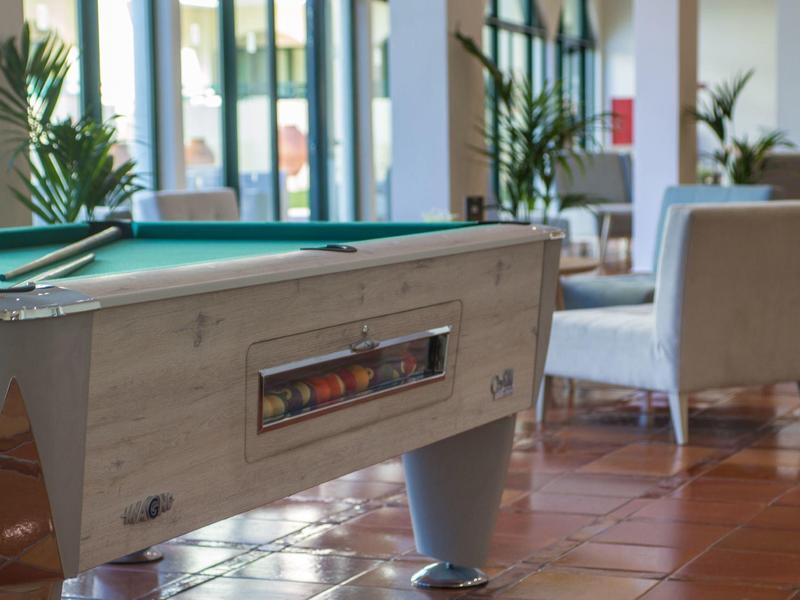 Billardtisch mit grüner Spielfläche in hellem Raum mit Terrakottaboden und Stühlen im Hintergrund.