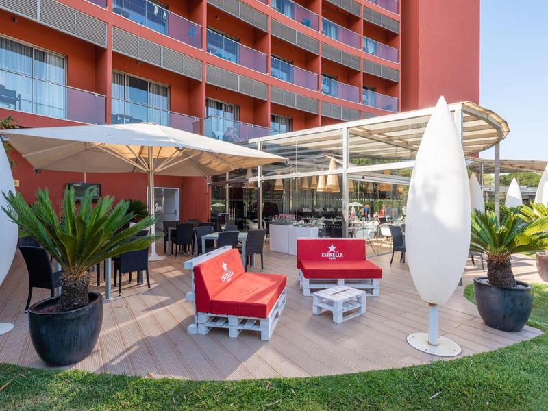 Terraza de hotel con muebles rojos, sombrillas y plantas delante de edificio de varios pisos.