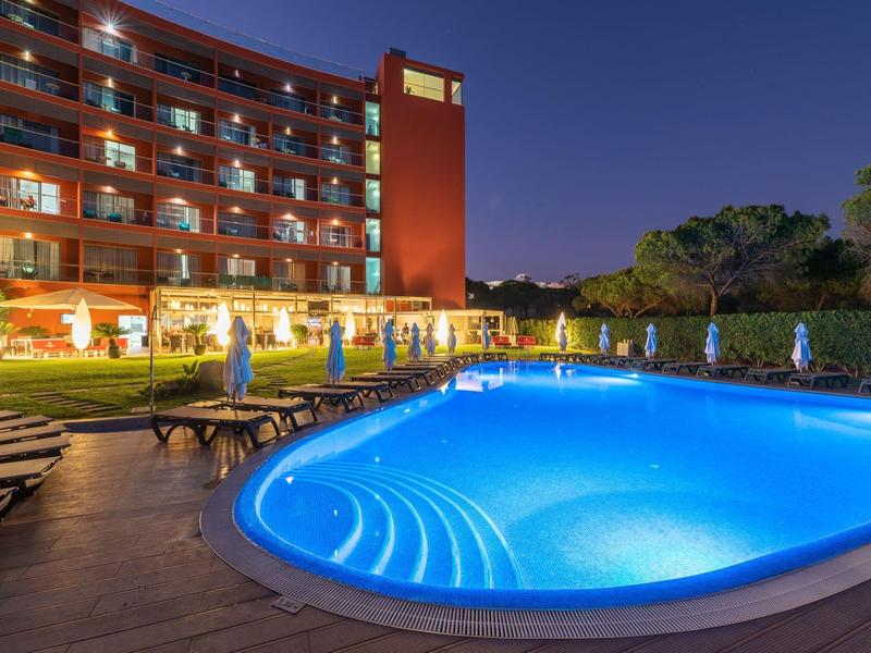 Piscina iluminada de noche frente a un edificio de hotel con hamacas y personas caminando