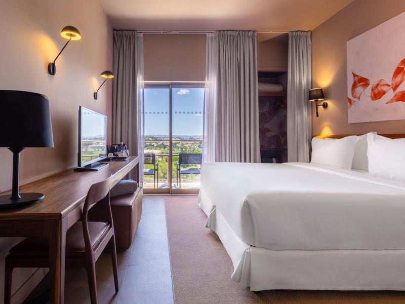 Habitación de hotel moderna con cama doble, escritorio y ventana grande con vista a paisaje verde.