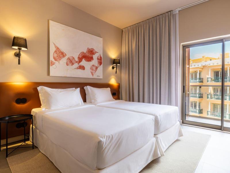 Habitación de hotel luminosa con dos camas, mesitas, lámparas de pared y balcón con vista.