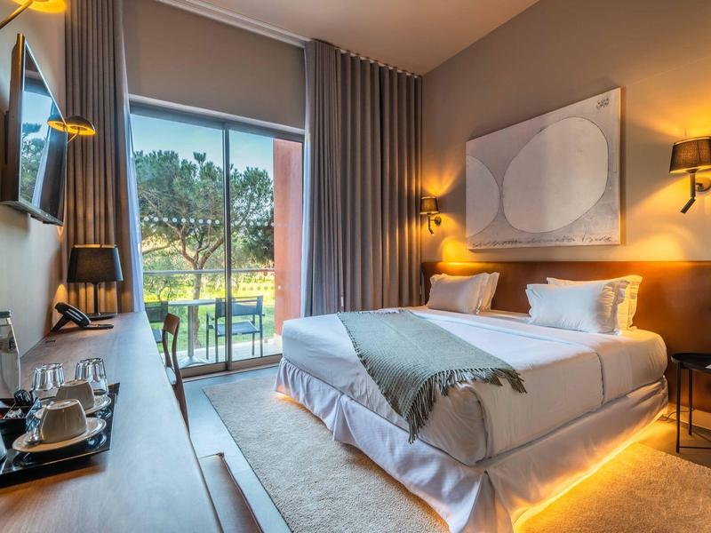 Habitación de hotel moderna con cama doble, gran ventana y vista a un jardín al atardecer.