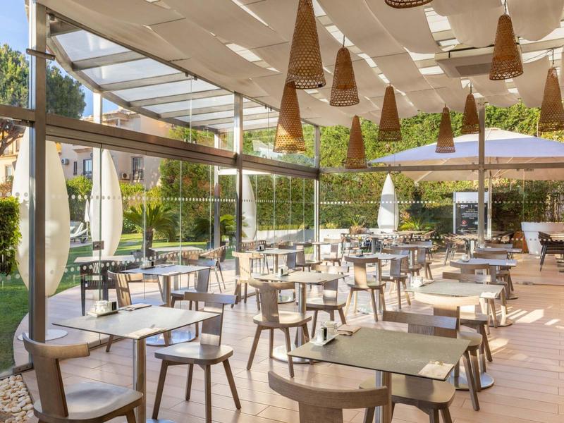 Área de comedor al aire libre en un hotel con mesas, sillas y sombrillas bajo un techo parcial.