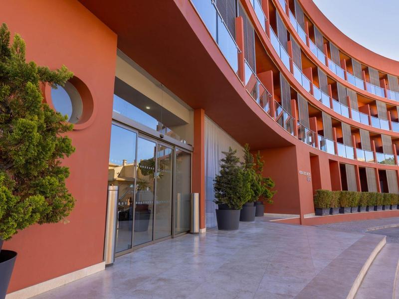 Fachada curva de hotel con entrada de cristal y plantas en macetas frente al edificio.