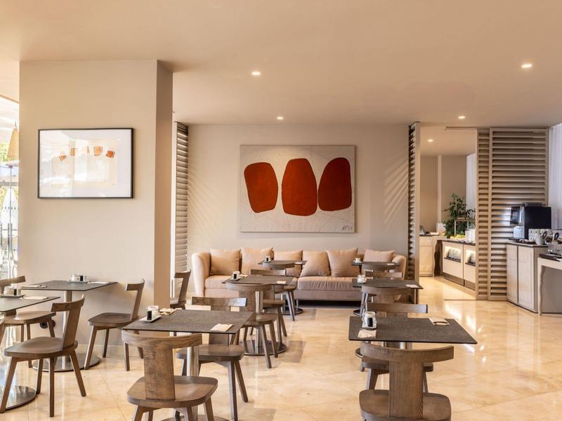 Sala de desayuno moderna con mesas, sillas y zona de sofá acogedora en ambiente claro.