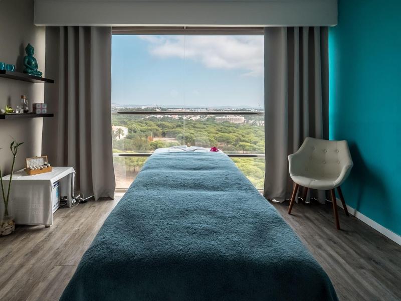 Sala de tratamiento con camilla de masaje y vista al paisaje a través de grandes ventanas.