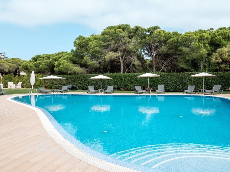 Piscina exterior redonda con agua azul, tumbonas y sombrillas en entorno verde.