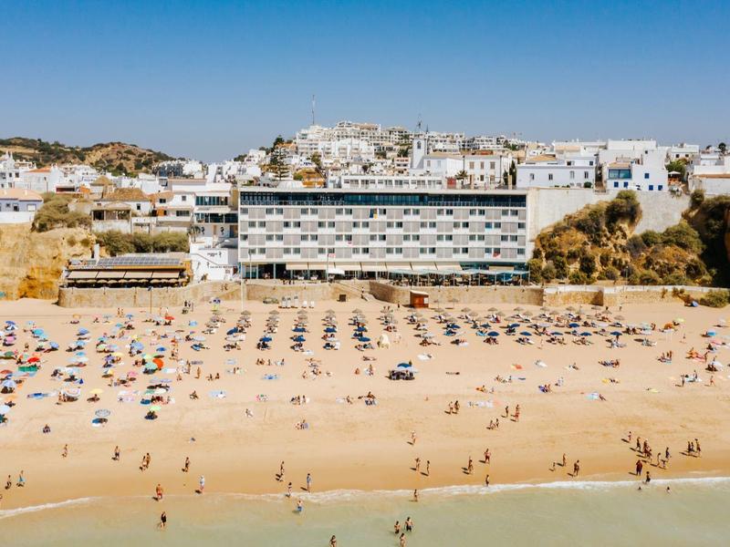 Piaszczysta plaża z wieloma parasolami, za nią duży hotel i domy na wzgórzu.