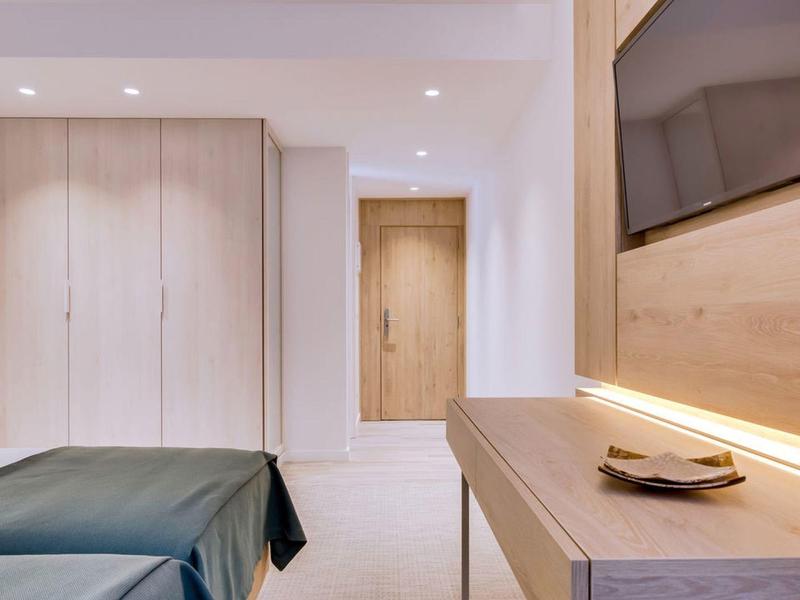 Jasny pokój hotelowy z podwójnym łóżkiem, szafą i telewizorem nad drewnianą półką ścienną.