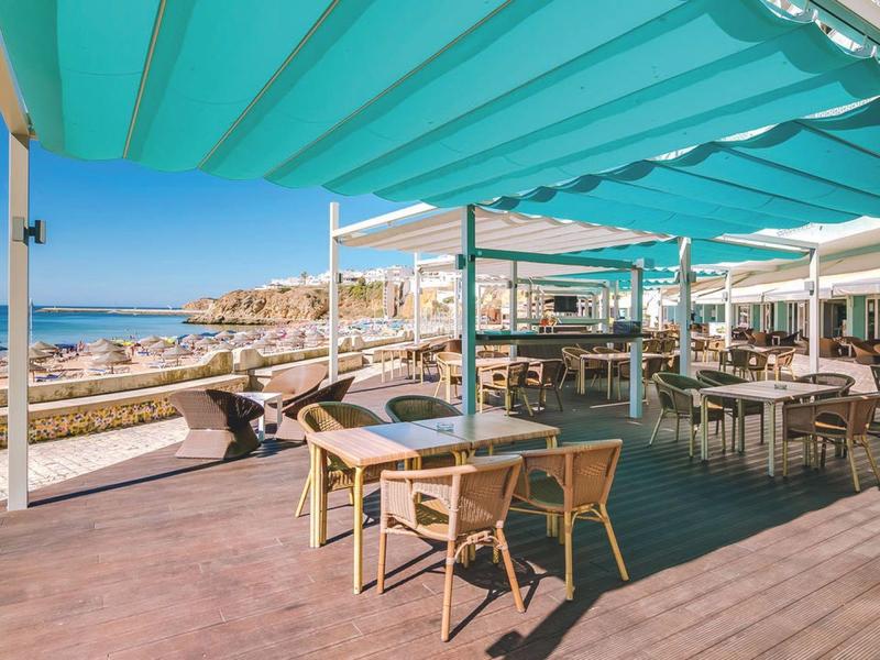Taras z stołami i krzesłami pod niebieskimi markizami z widokiem na morze.