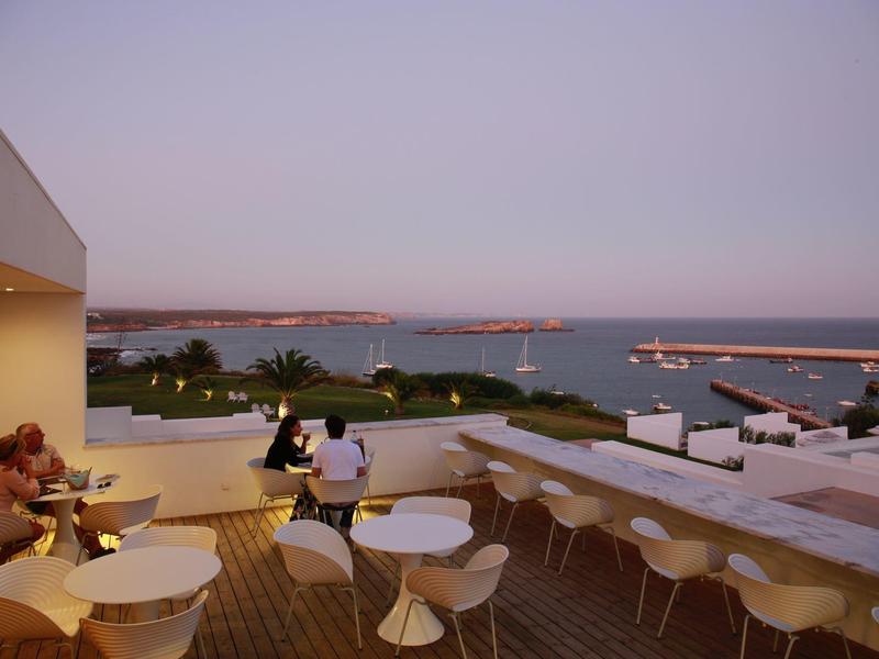 Terasa s bílými židlemi a stoly při západu slunce s výhledem na moře a přístav.