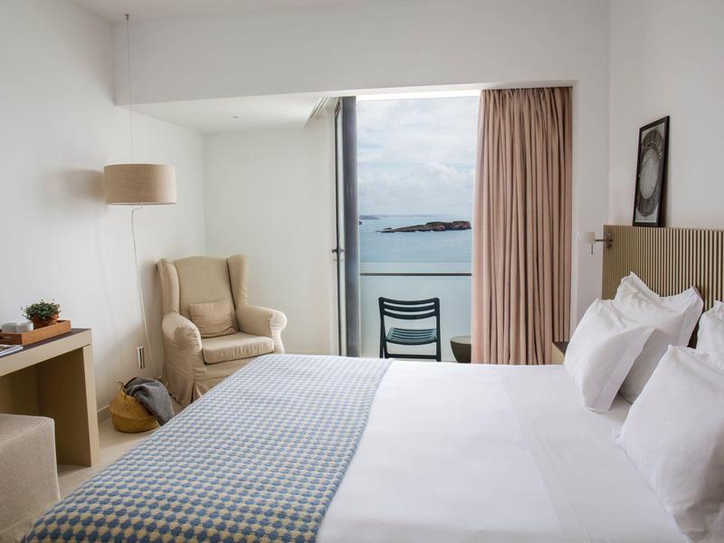 Světý hotelový pokoj s výhledem na moře, pohodlným křeslem a manželskou postelí s bílými polštáři.