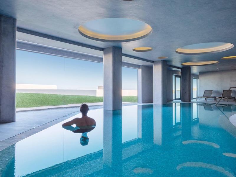 Uvolněný muž v krytém bazénu moderního hotelu s velkými okny a přirozeným světlem.