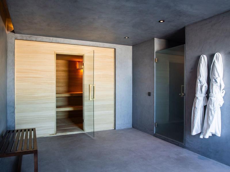 Moderní sauna s prosklenou sprchou, dřevěnou lavicí a bílými župany na zdi.