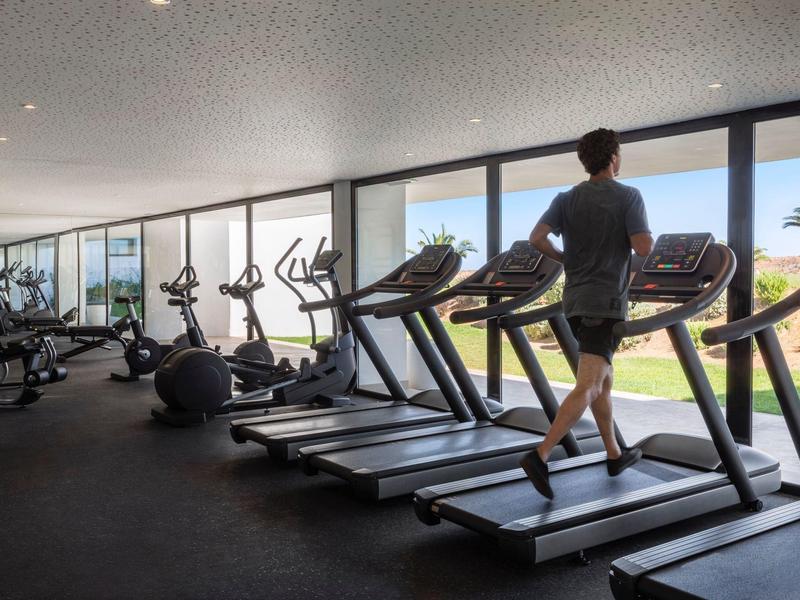 Osoba chodí na běžeckém pásu v posilovně s velkými okny a cvičebním vybavením.