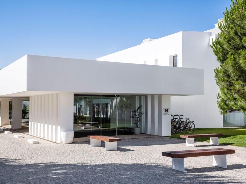 Moderní bílá budova s lavičkami a stromy pod jasnou oblohou