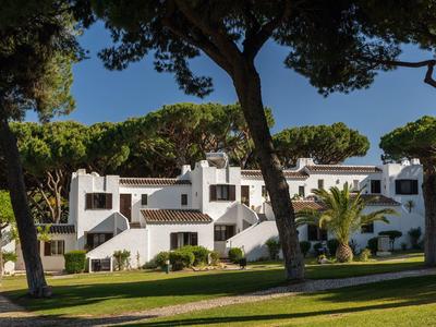 Weißes mediterranes Hotelgebäude mit blauen Akzenten und großen Bäumen im Vordergrund.