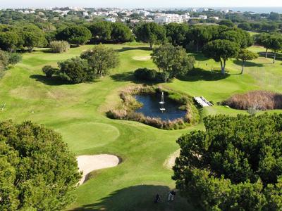 Blick auf einen grünen Golfplatz mit Teich, Bäumen und Sandbunkern unter blauem Himmel.