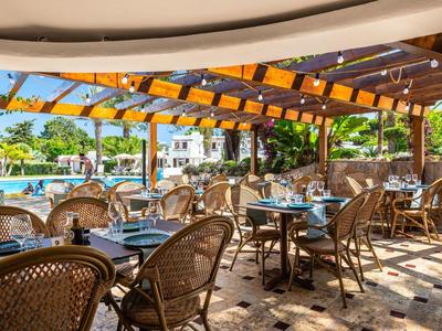 Freiluftrestaurant mit Korbstühlen und Tischen unter einer Holzpergola neben einem Pool.