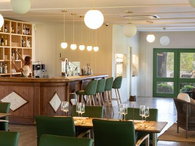 Helle Hotelbar mit Holztheke, grünen Sesseln und runden Lampen an der Decke.