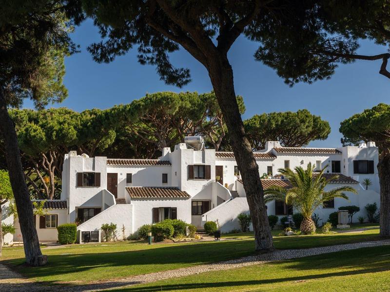 Weißes mediterranes Hotelgebäude mit blauen Akzenten und großen Bäumen im Vordergrund.