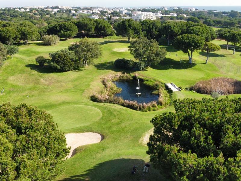 Blick auf einen grünen Golfplatz mit Teich, Bäumen und Sandbunkern unter blauem Himmel.