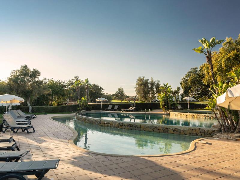 Ein ruhiger Hotelpool mit Liegestühlen und Sonnenschirmen umgeben von Bäumen bei Sonnenuntergang.