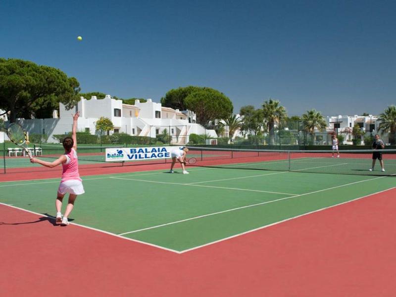 Zwei Frauen spielen Tennis auf einem roten und grünen Außenplatz bei sonnigem Wetter.