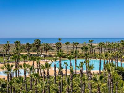 Großer blauer Pool umgeben von Palmen und grünen Pflanzen unter klarem blauem Himmel am Meer.