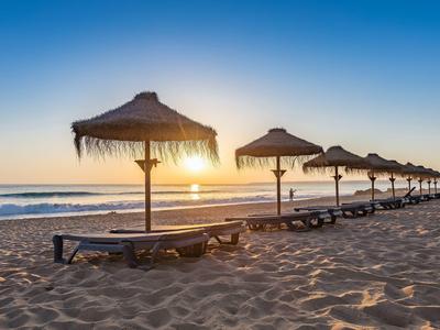 Strand mit mehreren Sonnenliegen und Schirmen aus Stroh bei Sonnenaufgang über ruhigem Meer.