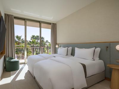 Helles Hotelzimmer mit Doppelbett, großen Fenstern und Blick auf Palmen im Außenbereich.