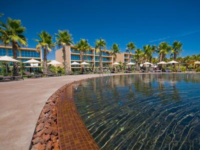 Klares Wasser vor einer Poolanlage mit Palmen und Sonnenschirmen unter blauem Himmel.