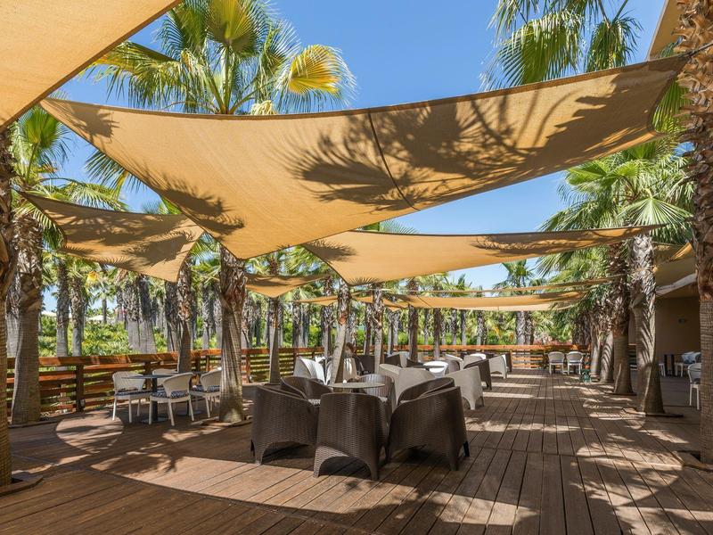 Outdoor-Restaurant mit hölzernem Boden, Tischen, Stühlen und beige Segeltüchern als Sonnenschutz.