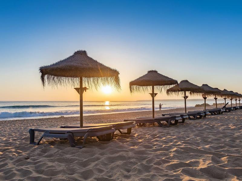 Strand mit mehreren Sonnenliegen und Schirmen aus Stroh bei Sonnenaufgang über ruhigem Meer.