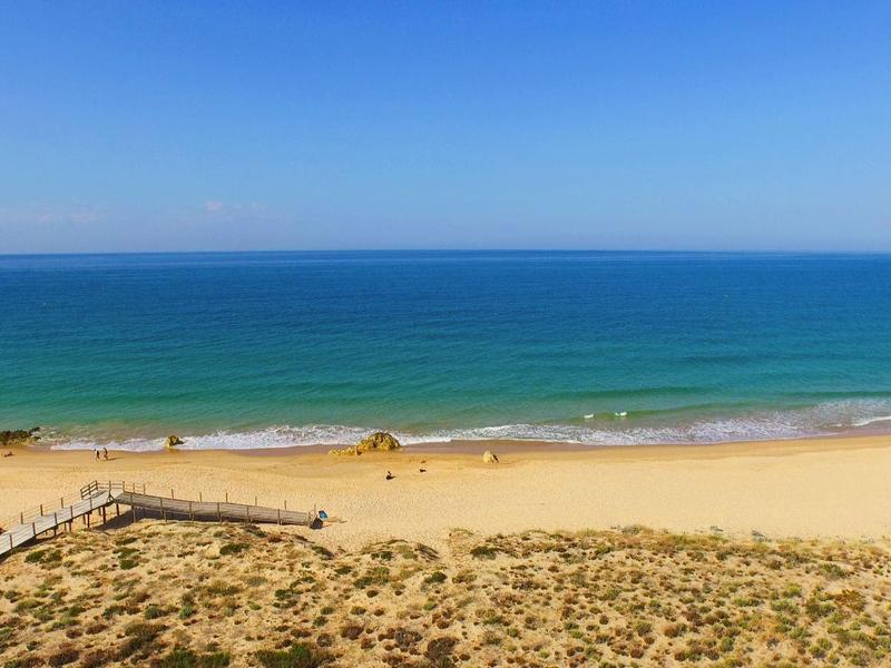 Breiter Sandstrand mit klarem blauem Himmel und türkisfarbenem Meer, Dünen im Vordergrund.