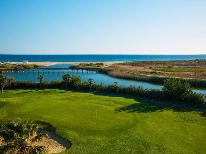 Grüner Golfplatz mit Sandbunker, Wasserhindernis und blauer Meereslinie im Hintergrund.