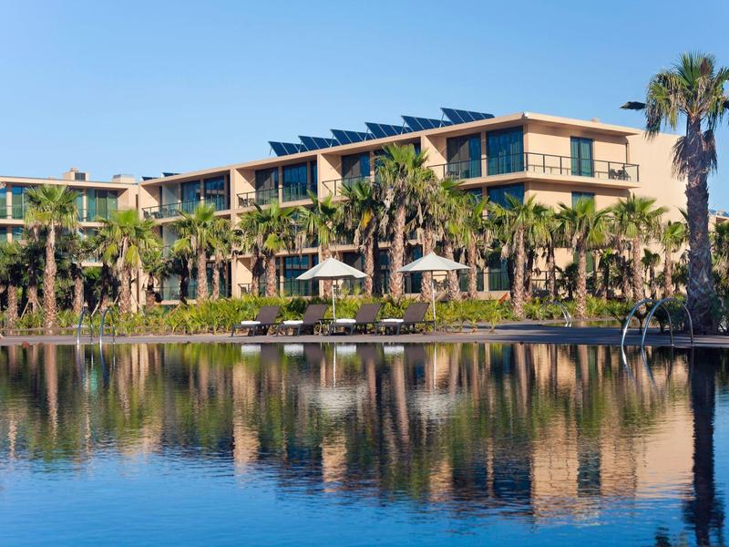 Helles, modernes Schlafzimmer mit Doppelbett, Tapetenmuster, Balkon und Blick auf Palmen.