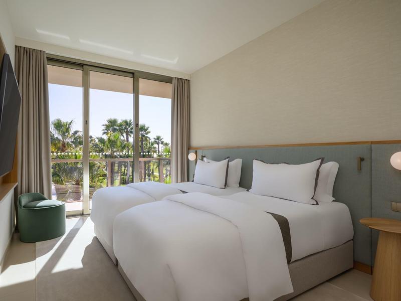 Helles Hotelzimmer mit Doppelbett, großen Fenstern und Blick auf Palmen im Außenbereich.