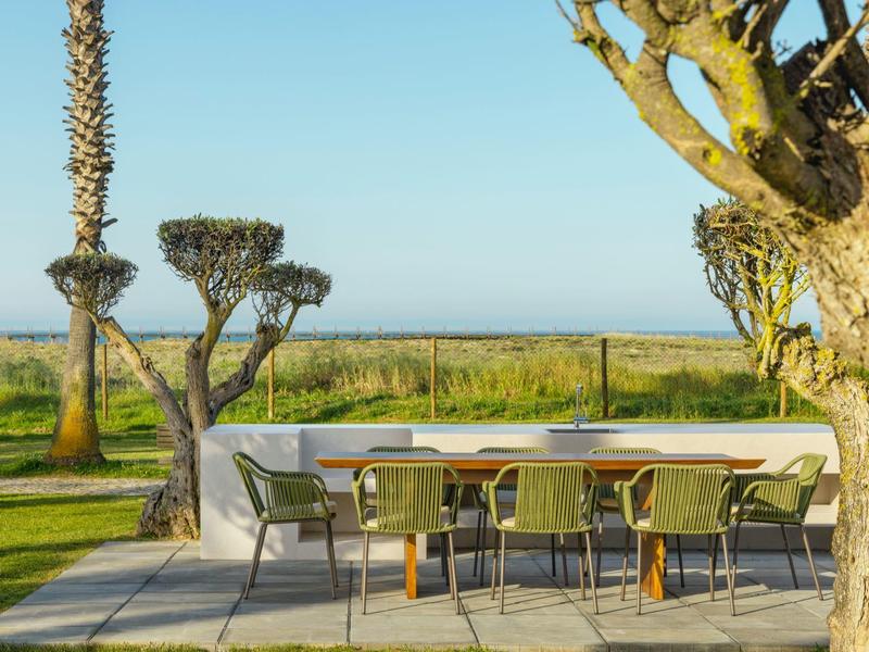 Moderner Gartentisch mit Stühlen auf Terrasse, umgeben von Bäumen, Wiese und blauem Himmel.