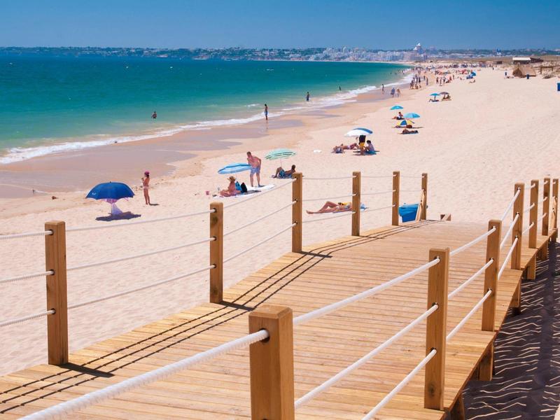 Strand mit hellem Sand, blauem Meer, bunten Sonnenschirmen und Holzsteg am sonnigen Tag.
