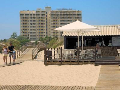 Strand mit Sand, Holzsteg, zwei Personen und einem Gebäude im Hintergrund bei sonnigem Wetter.