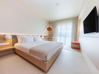 Helles, modernes Hotelzimmer mit Doppelbett, Wandfernseher und Balkon mit Vorhängen.