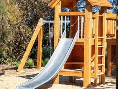 Holzspielplatz mit Rutsche in einem sonnigen Außenbereich, umgeben von grünen Bäumen.