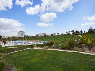 Zelená parková plocha se stromy před hotelovým komplexem pod modrou oblohou s mraky.