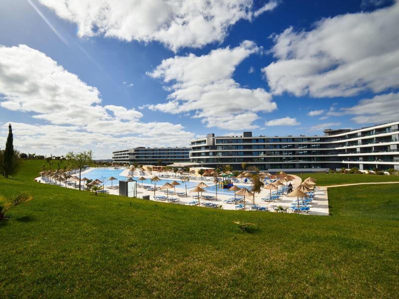 Moderní hotel s velkým venkovním bazénem a lehátky, obklopený zeleným trávníkem a modrou oblohou.