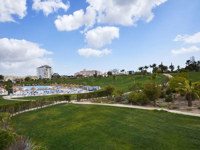 Zelená parková plocha se stromy před hotelovým komplexem pod modrou oblohou s mraky.