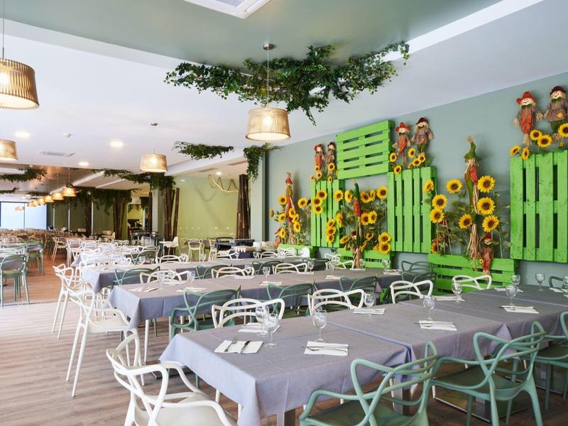 Moderní jídelna se šedými stoly, bílými židlemi a zelenými dekoracemi se slunečnicemi.