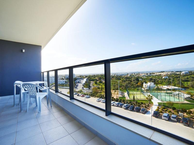 Balkon se skleněným zábradlím, bílým stolem a židlemi s výhledem na park a město.